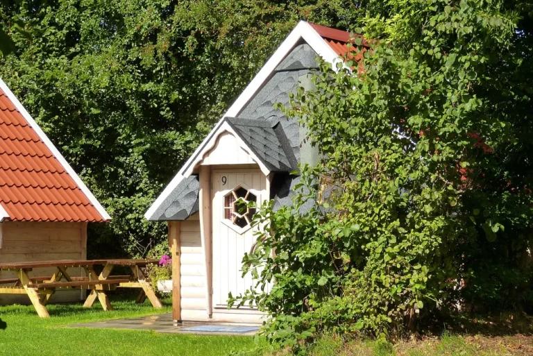 Een sfeervol, wit houten huisje met nummer 9 en een zeshoekig raampje, gelegen in een groene omgeving naast een picknicktafel onder een rood dak.